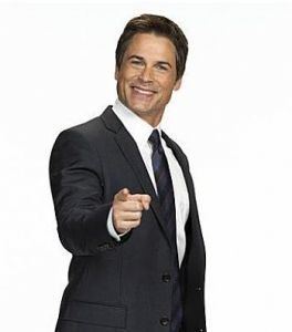 A photo of Rob Lowe in a jacket and tie smiling and pointing directly at the photographer