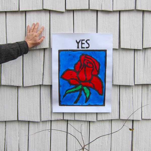 Photo of block-print poster of a red rose on a blue background with the word "YES" above, against the background of a grey clapboard wall