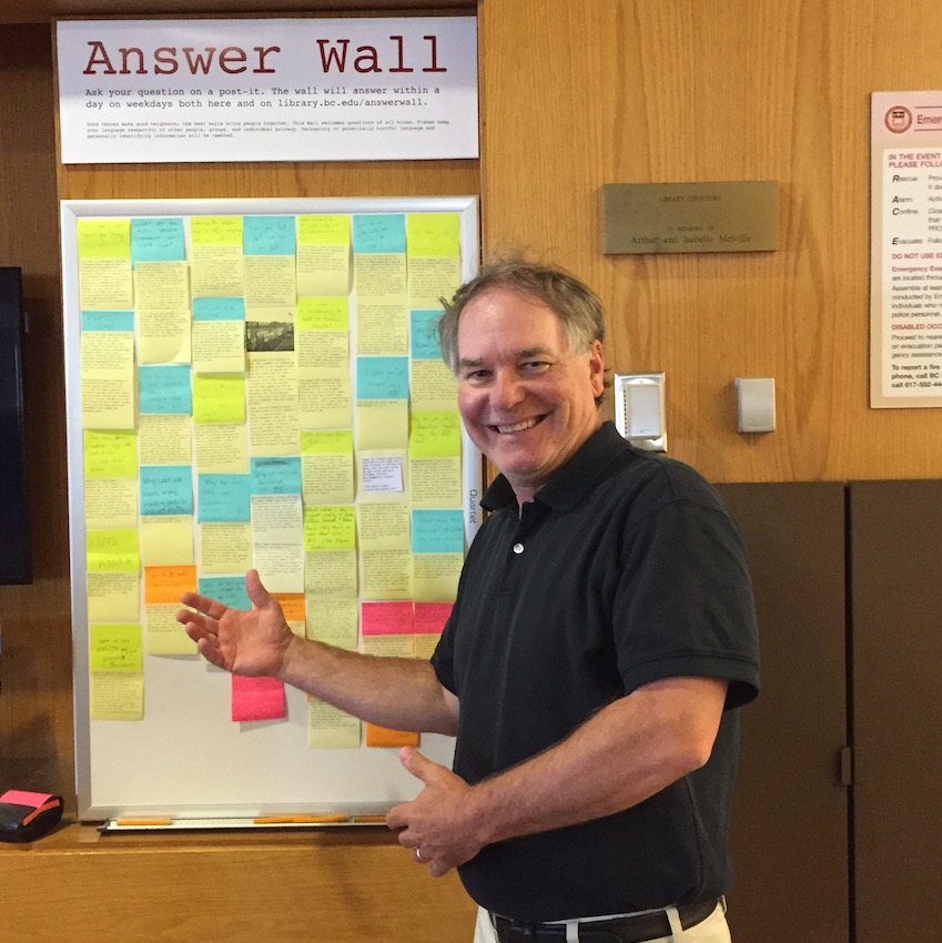 BC University Librarian Tom Wall posing, smiling, in front of the Answer Wall in the O'Neill Library Lobby