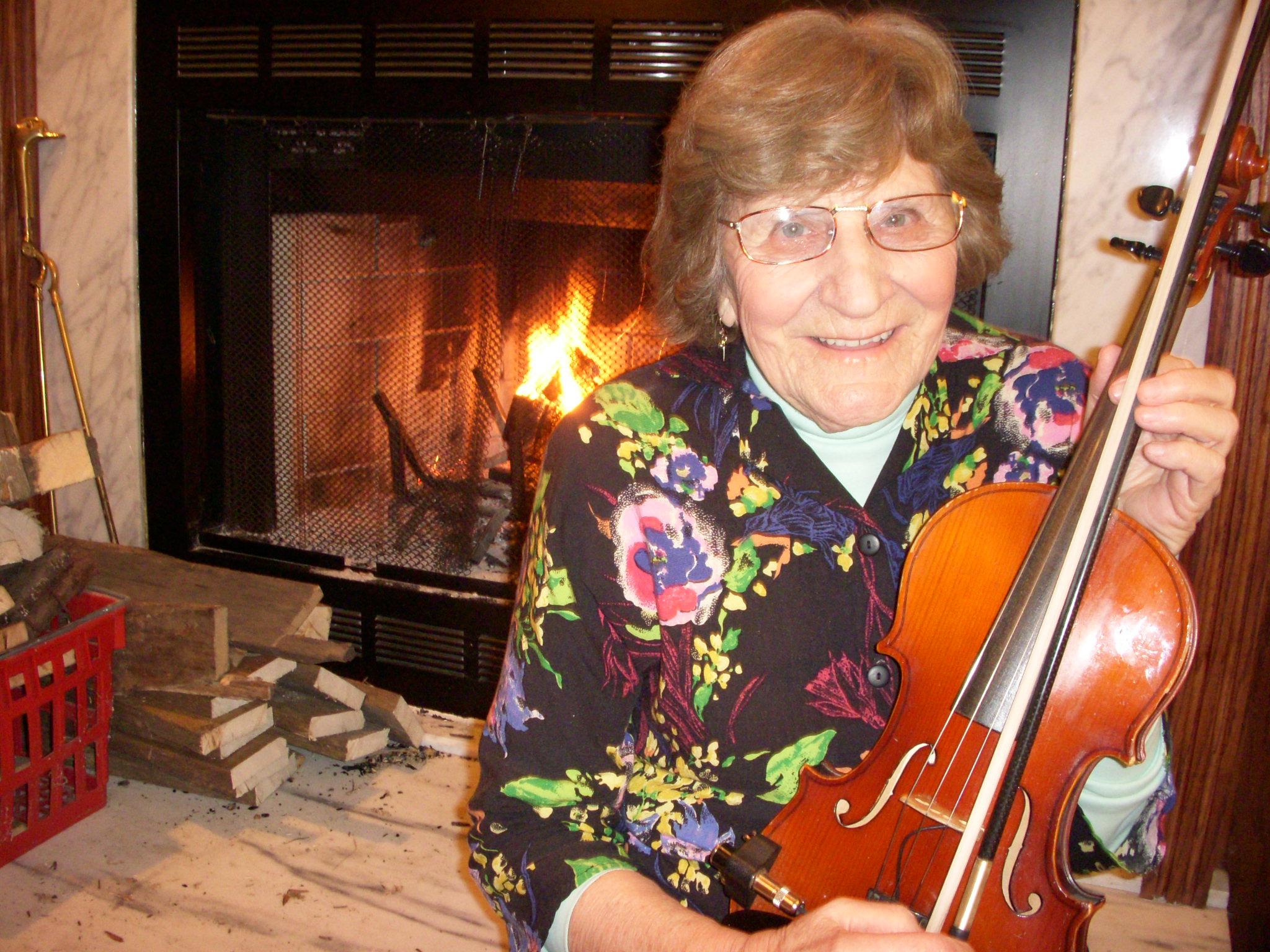Photograph of Matilda Murdoch, sitting with a violin in front of a lit fireplace