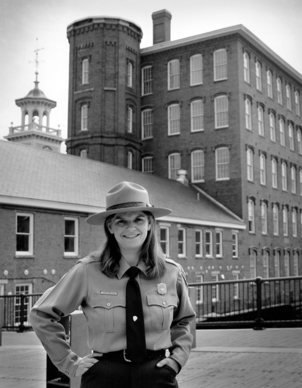 Photograph of Chrysandra 'Sandy' Walter in Park Ranger attire, standing in front of an old mill in Lowell, Mass.