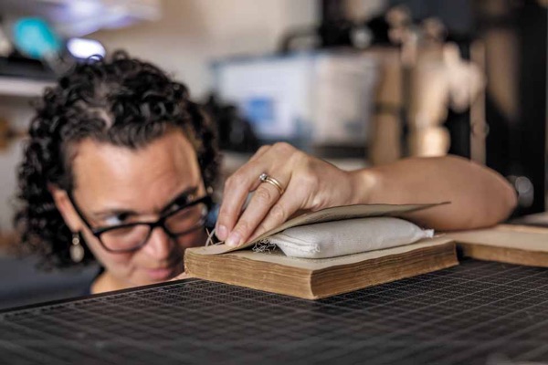 A woman with dark curly hair peers over her black-framed glasses, out of focus, level with a book in the foreground that her left hand presses on the edge of a table. A needle pokes upward past the book’s binding.