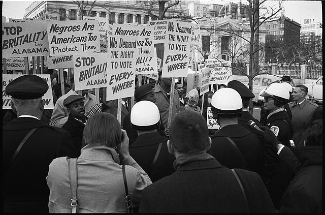 Black and white image of a tight cluster of African American demonstrators outside the White House, with signs "We demand the right to vote, everywhere" and signs protesting police brutality against civil rights demonstrators in Selma, Alabama