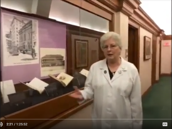 slightly blurry zoom capture image of Barbara Adams Hebard in white conservator lab coat gesturing next to a glass display case of books