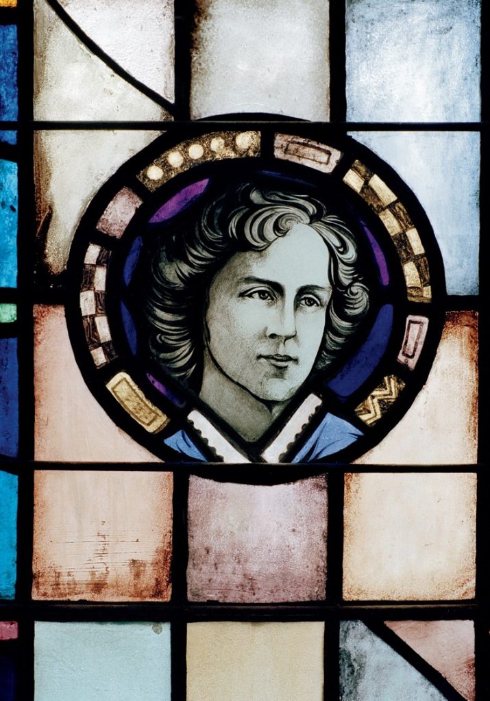 A stained glass portrait of Guiney's head surrounded by a purple and black patterned circle, with blue, red, and yellow square windowpanes around it.