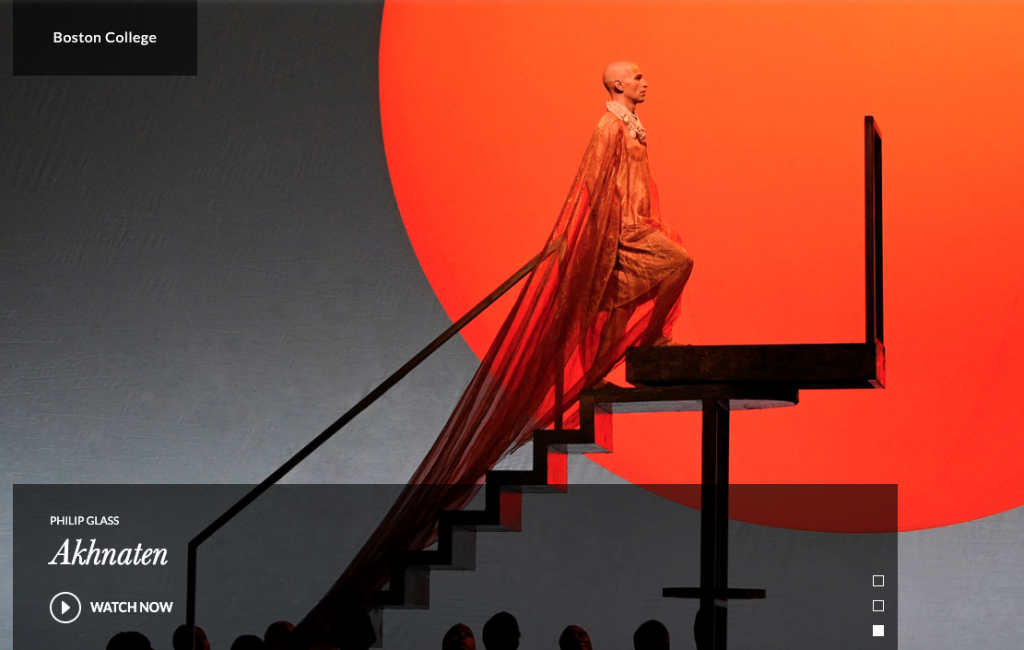 Dramatic image of character in an orange costume with a long train climbing stairs against a background of a large stylized orange sun in the Met staged opera of Akhnaten.