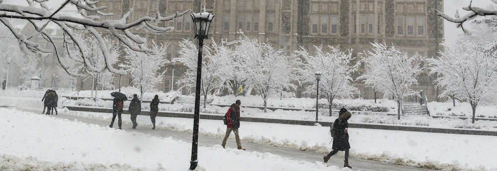 Silhouettes of people walking across the O'Neill Library quad at Boston College in the snow. Small trees are outlined in snow in front of Gasson Hall.