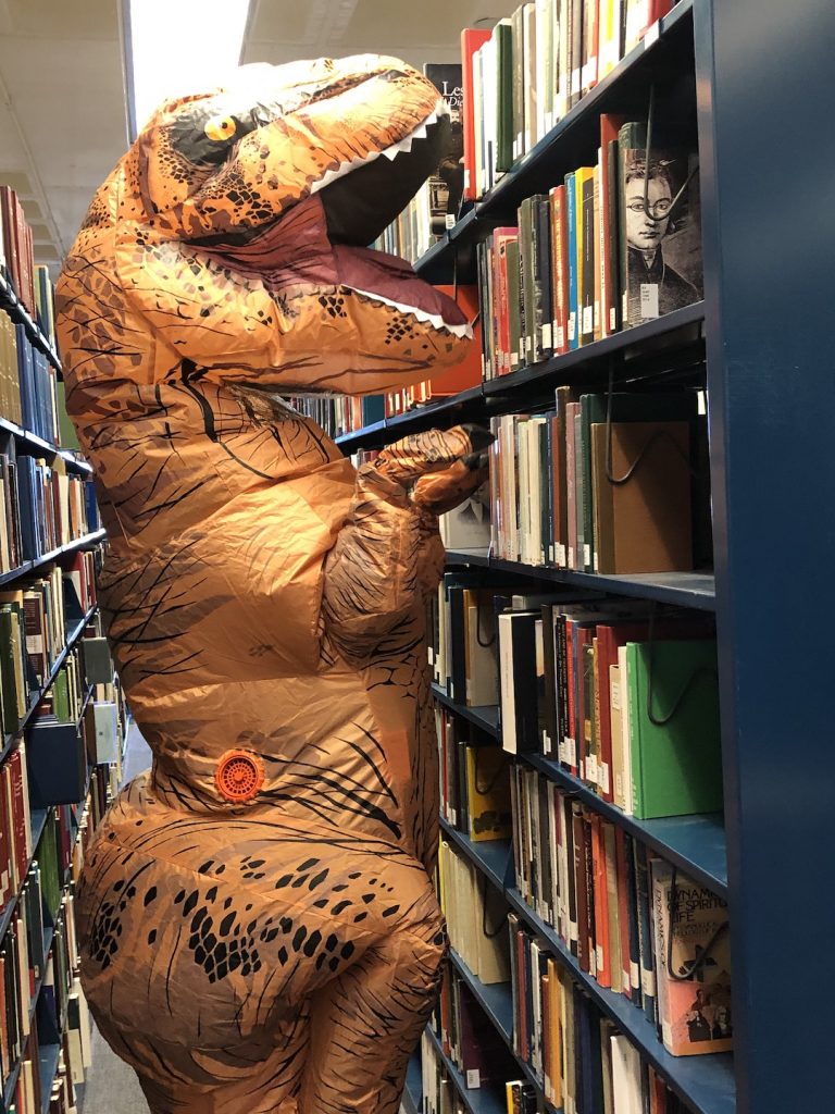 Person in inflatable T-Rex costume between two rows of tall library shelving full of books.