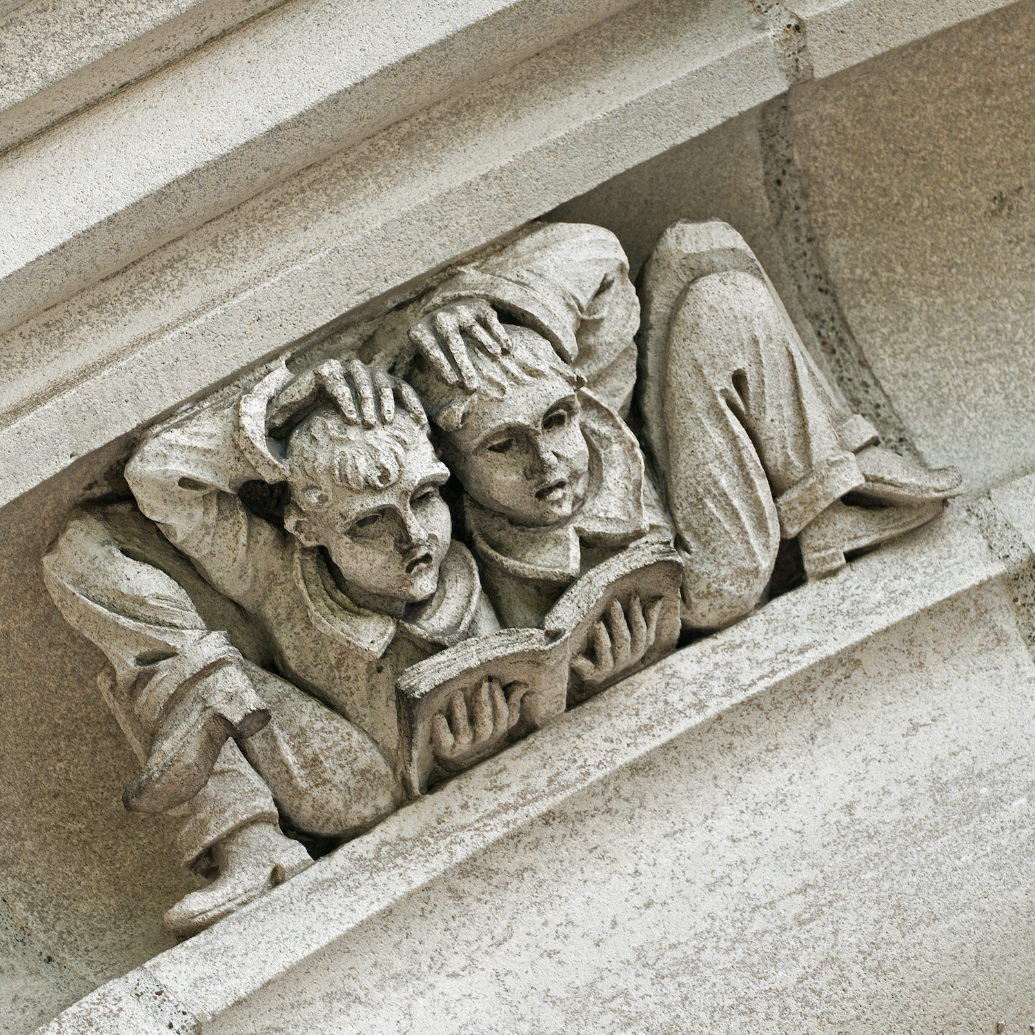 Detail of a whimsical gargoyle-like stonework figure of two students resting back to back while reading, and holding their heads in confusion.