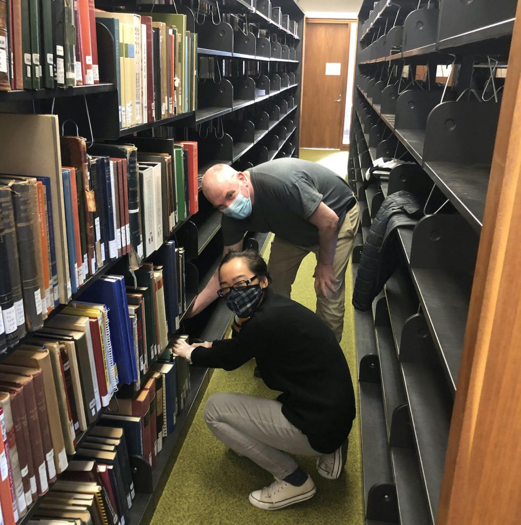 A man and a young woman crouch in a narrow library passage between shelves, both touching a book on the bottom shelf. Ranks of empty shelves are behind them. Both are wearing COVID pandemic masks.