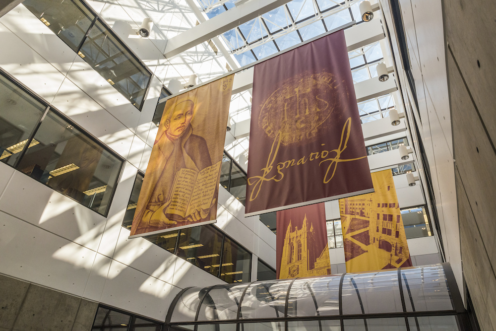 Bright morning sun streams through a large skylight into an atrium with red and yellow banners featuring St. Ignatius and Bapst and O'Neill Libraries.