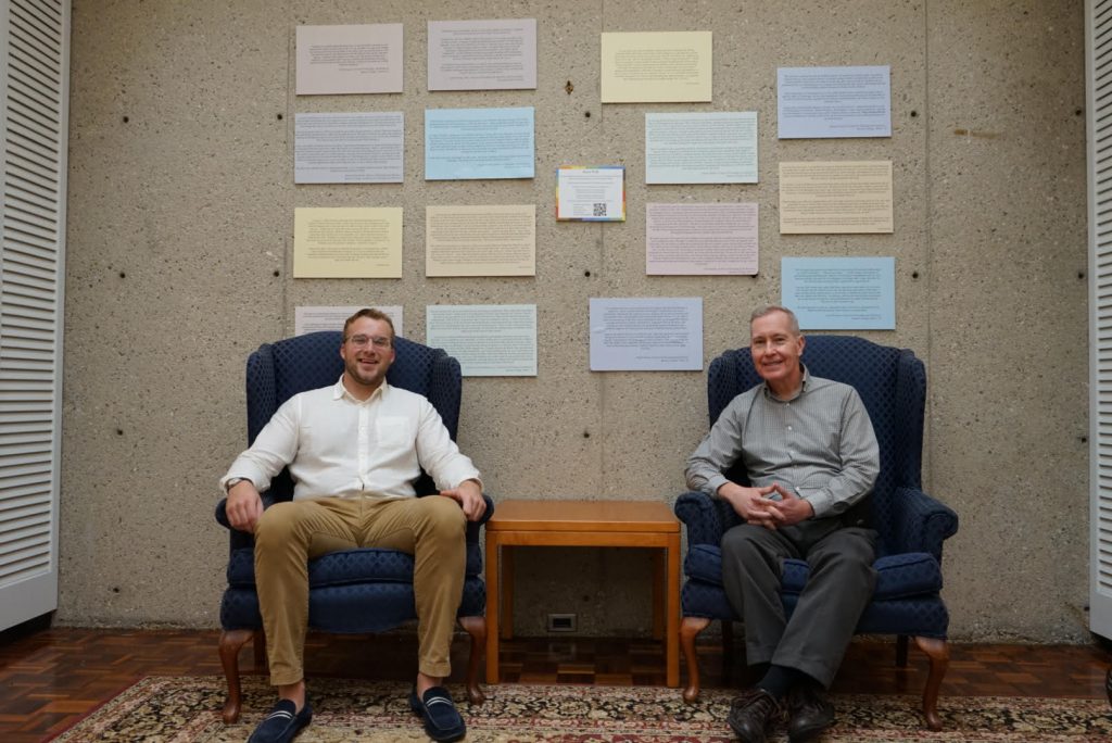 Two men in dark blue armchairs sit facing the camera; behind them are colored squares of paper covered in small print, mounted on a concrete wall.