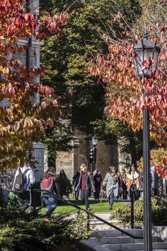 A few dozen students in light jackets under orange and green leaves in Fall on the BC campus, with stairs in the foreground