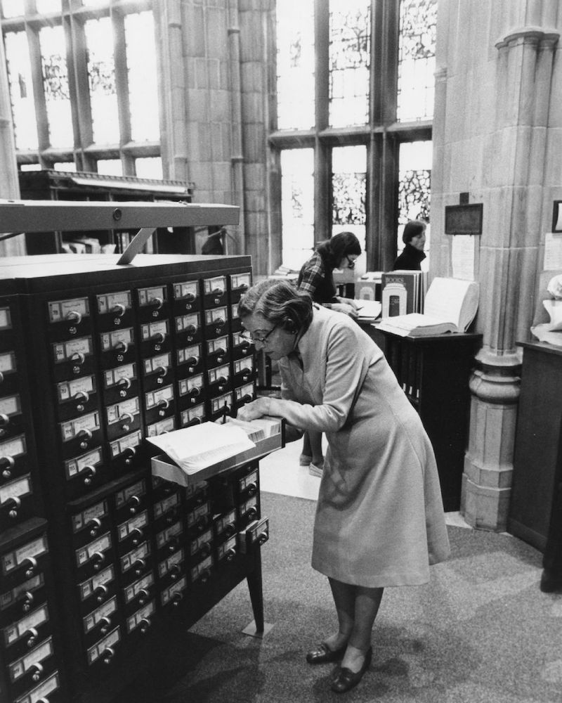 Black and white photo of an elderly librarian in a long-sleeved dress peers through her glasses into the contents of a card catalog drawer. A pencil pokes out of the cards, marking a location.