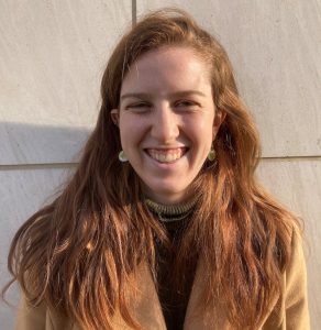 Close-up photograph of person with long red hair and earrings, in a tan jacket, smiling broadly, in late afternoon sun.