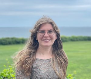 Photo of person with long hair, round glasses, and multicolored knit top, with a background of grass, ocean, and sky