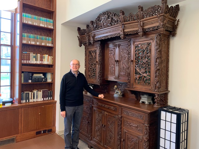 A man in grey slacks and a black windbreaker stands with one hand resting on the desk surface of a wooden cabinet with ornate carvings of scenes of nature and animals.