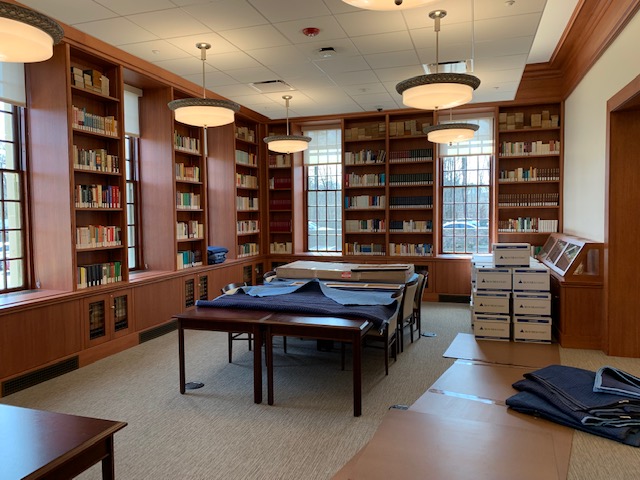 Nicely finished dark wooden shelves line a room with wooden tables, tall windows, and hanging light fixtures. Moving blankets cover the tables and stacks of boxes sit in the far corner