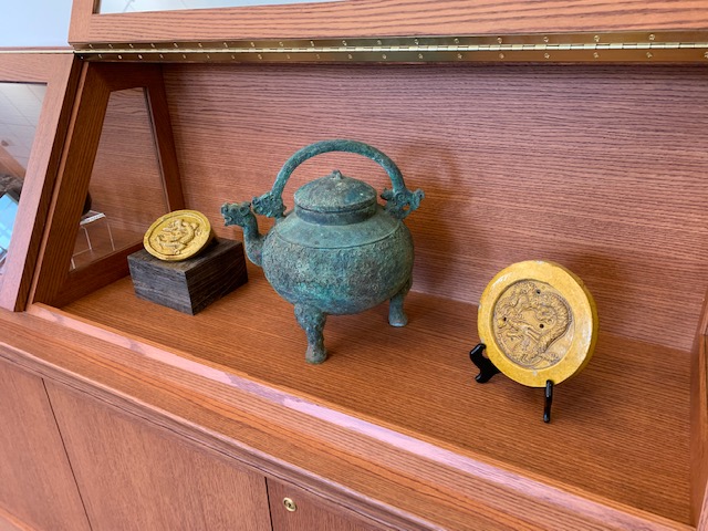 A bronze teapot with a green patina between two yellow glazed ceramic disks with bas-relief dragons in a wooden display case