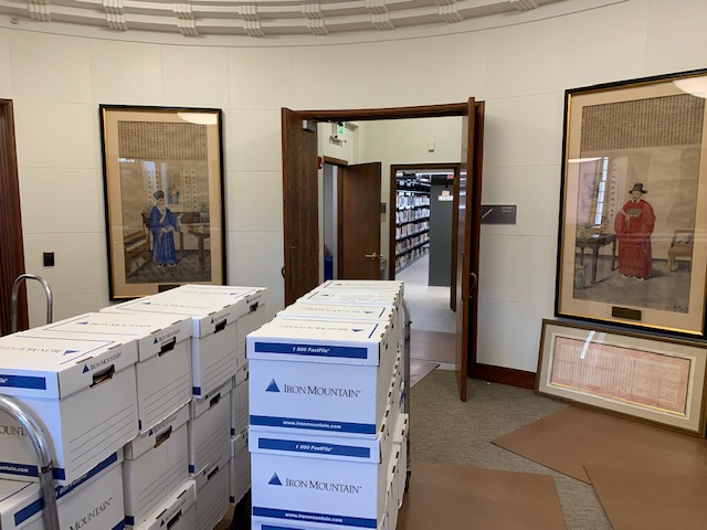 A few dozen boxes are in the foreground, while in the background two Chinese paintings lean on the wall; through a door empty metal shelving is visible