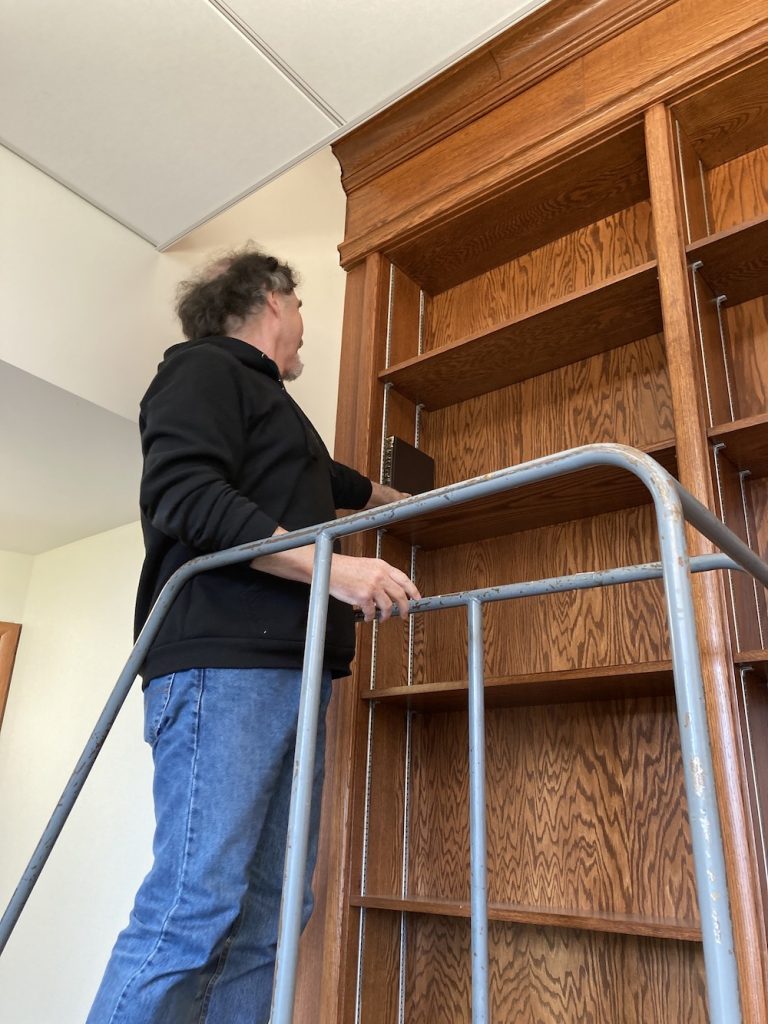 A man in jeans and a black sweatshirt stands on a stepladder while placing a book on nicely finished but empty wooden shelves.