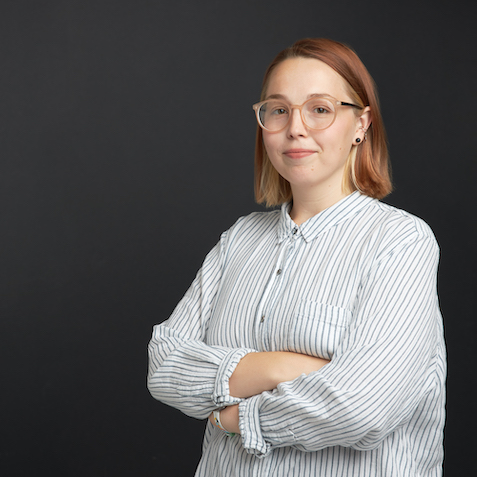 3/4 profile portrait of a person with shoulder length red hair, black earrings, round-framed glasses, and a white shirt with vertical blue stripes, posed against a dark gray background