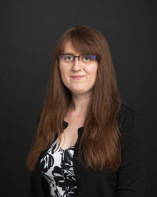 Photo portrait of person with long brown hair in black and white outfit, with glasses, against a dark background