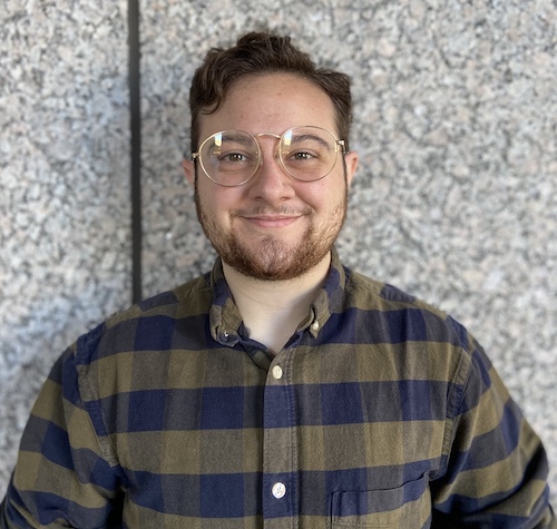 Photo portrait of person facing camera with glasses, short brown hair, and beard, in green and blue striped shirt against a granite wall background