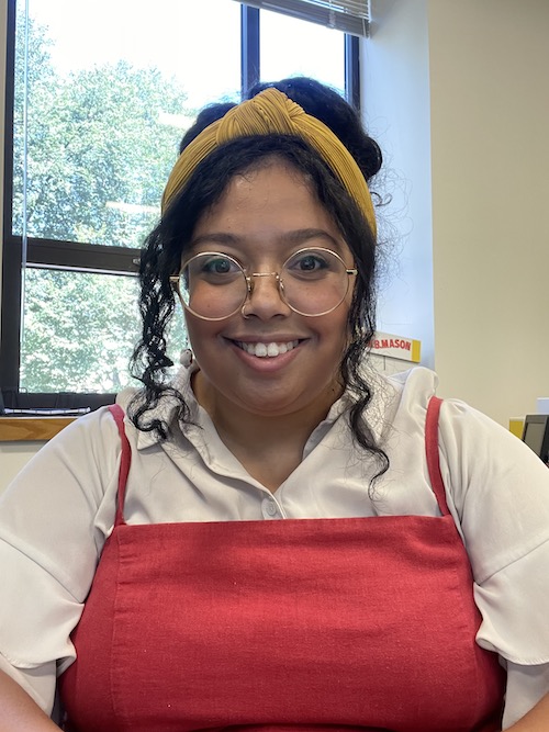 Photo portrait of a smiling person with dark hair in a yellow head-wrap, white top, and red dress.