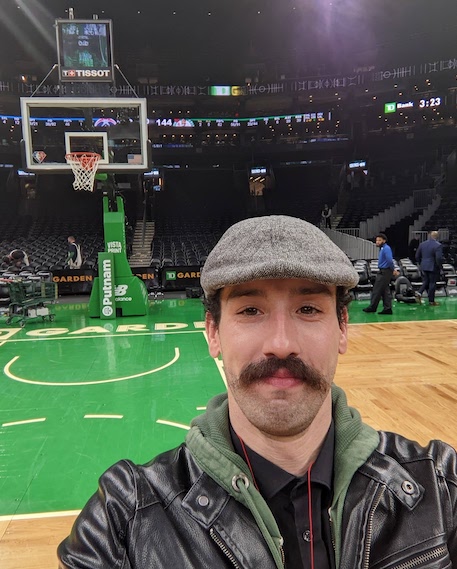 A smiling person with short brown hair, a gray wool cap, a moustache, green hoodie, and brown leather jacket in front of a background of a basketball court in a stadium