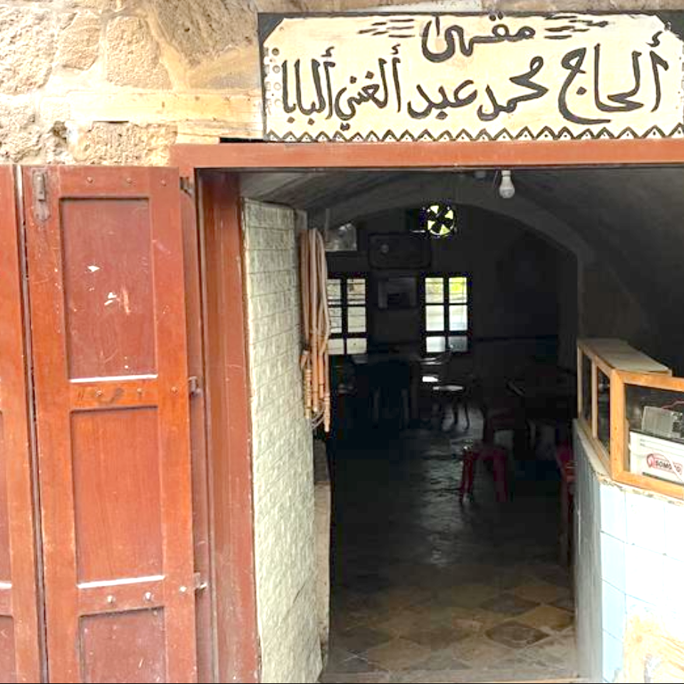 Open wooden door on a stone building revealing a shadowy interior with tiled floor and cafe tables. Above the door is an Arabic inscription.