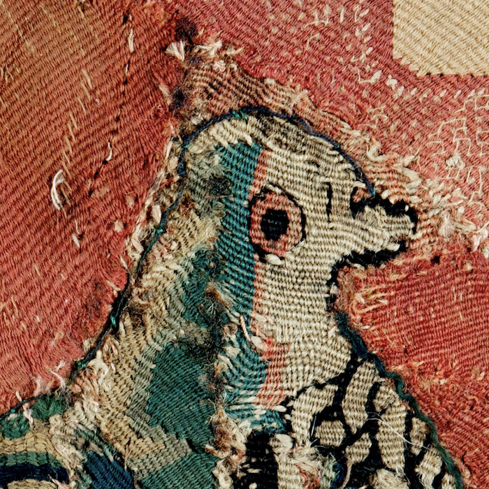 Close-up of antique needlework in blue, red, and black of a dove on a red background.