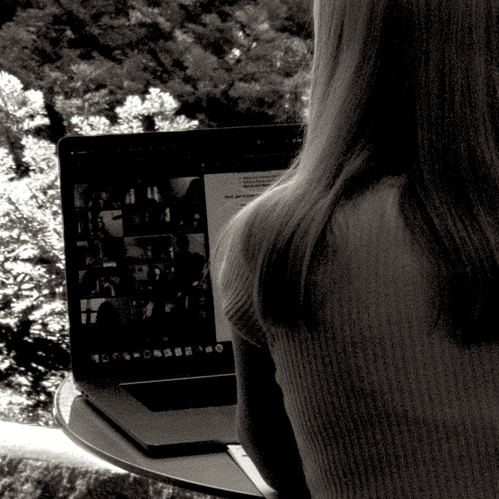 the blonde hair draped on the back of a shoulder of a person looking at a laptop screen, against a background of leaves in bright sunshine