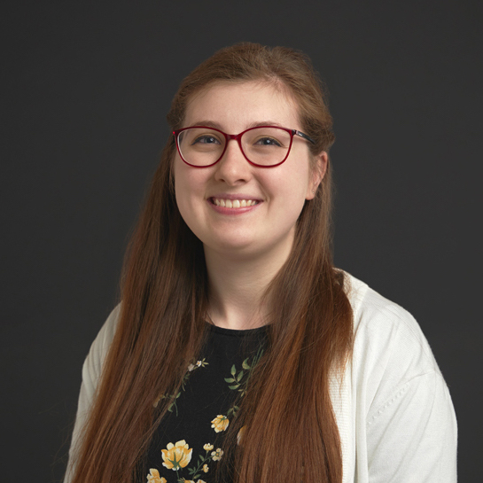 Photo of smiling person with red glasses and long brown hair, in a black and yellow floral shirt and white sweater against a dark background.