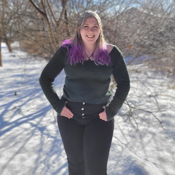 Photo portrait of a person with long blonde hair with purple ends, in a dark sweater and black jeans against a snowy, sunny background