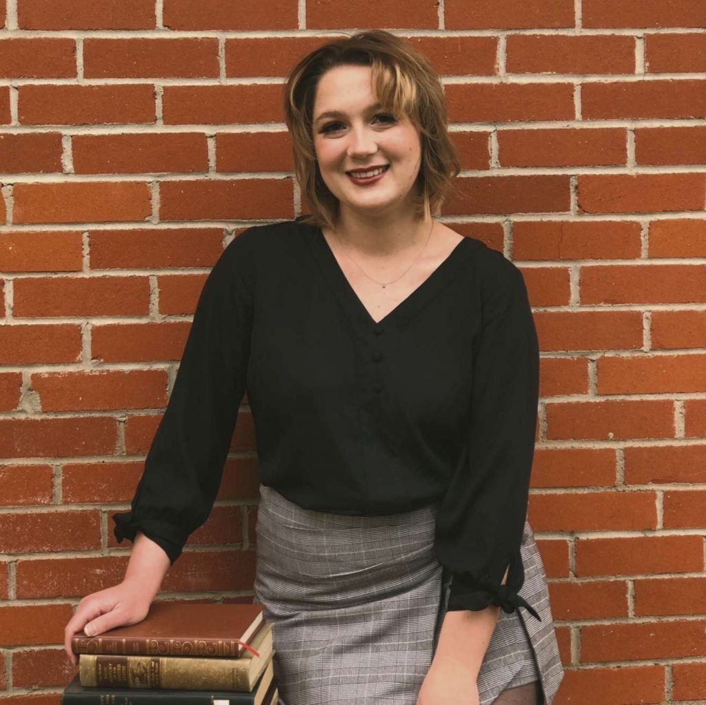 Photo portrait of a blonde person in v-neck black top and grey skirt, one hand on a stack of books, against a background of bricks.