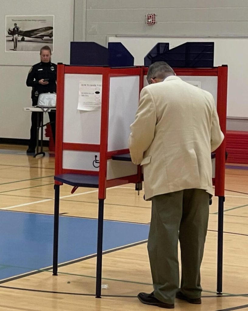 A man with his back to us at a voting booth in a gym with a police officer in the background.
