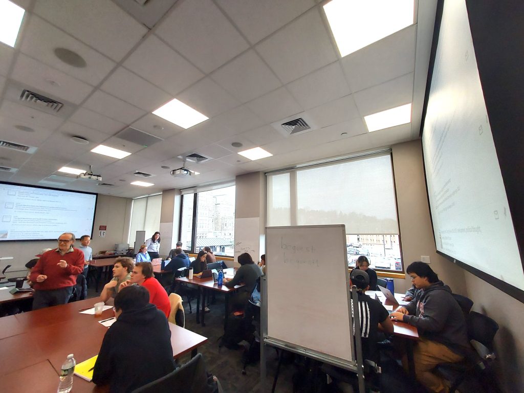 A wide angle shot of a busy classroom with students working and talking to each other at tables, and two projection screens