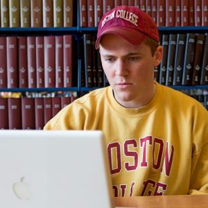 A person in a red Boston College baseball cap and yellow Boston College sweatshirt intently stares at a laptop computer screen in front of a background shelves-ful of books.