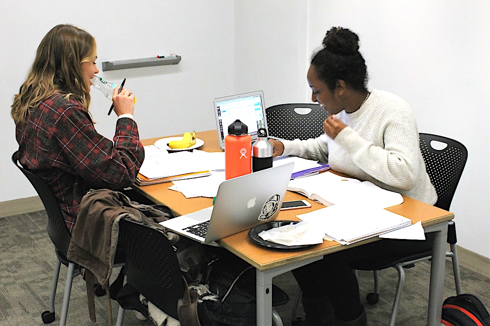 Two people seated on opposite sides of a small table crowded with paper, laptops, water bottles, and snacks, laugh together, in front of a background of white walls.