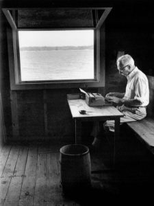 In a black and white photo, a white haired man, seen in profile, sits at a simple table with a typewriter next to an open square window looking out at a body of water. The mood is simple and rustic.