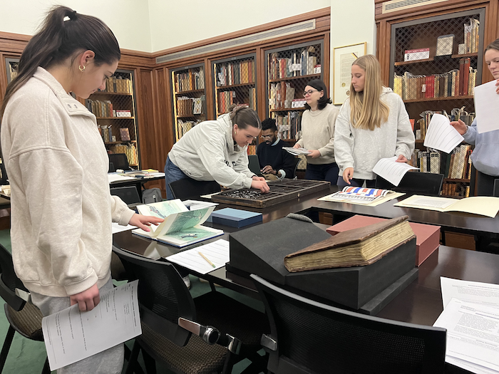 5 people examine various books and other objects on tables in a room surrounded by elegant wooden bookshelves secured by wire mesh doors.