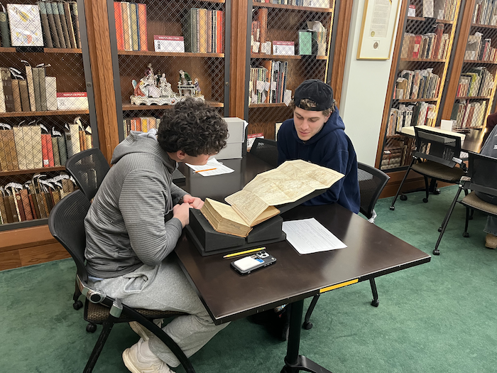 Two seated students on opposite sides of a table huddle over an old book with an interesting fold-out page, with full bookshelves in the background