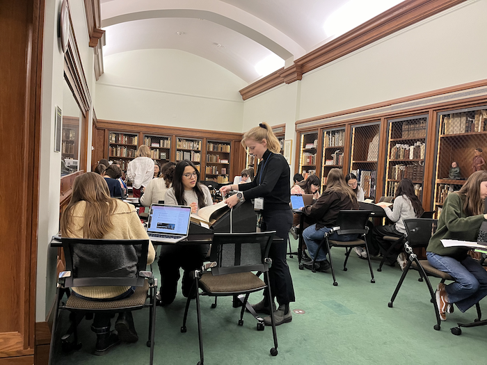 In an elegant room with an arched ceiling and walls lined with wooden shelving, a little over a dozen students sit and stand at tables intently examining books and using laptops.