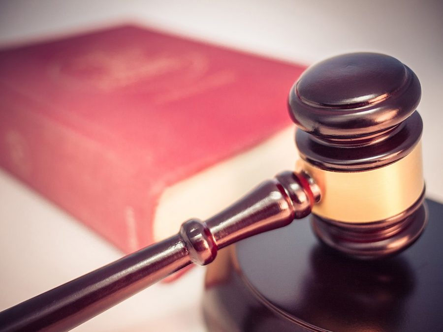 A wooden gavel with a brass center rests on a dark wooden disk, with an out of focus red hardcover book in the background