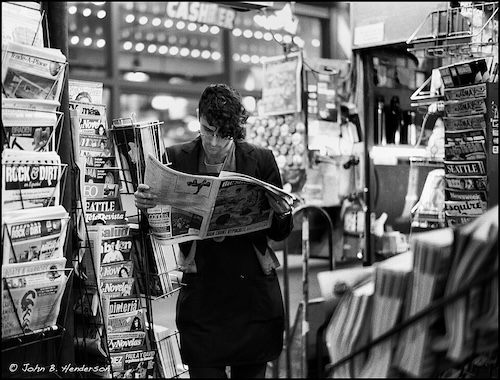 A person with classes and long dark wavy hair wearing a dark blazer stands reading a newspaper in a newsstand crowded with magazines and newspapers. The image is black and white.